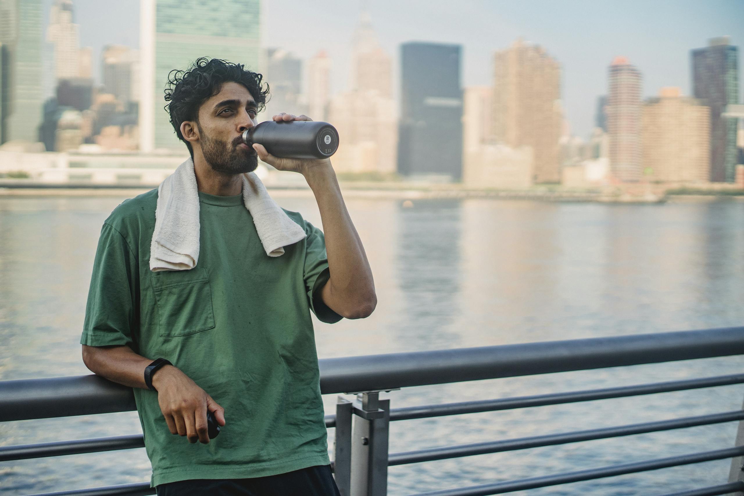 A man drinks water from a jug while leaning on a railing with a city skyline in the background.