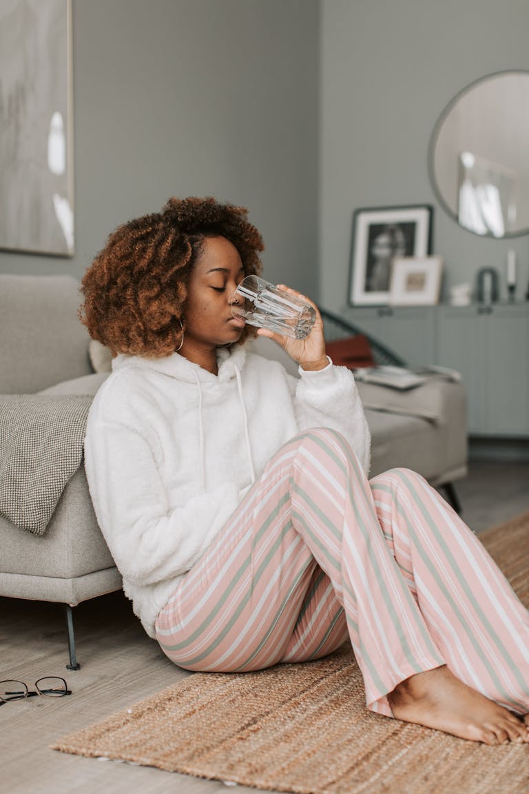 African American woman in hoodie sipping water, sitting on the floor in a cozy home environment.