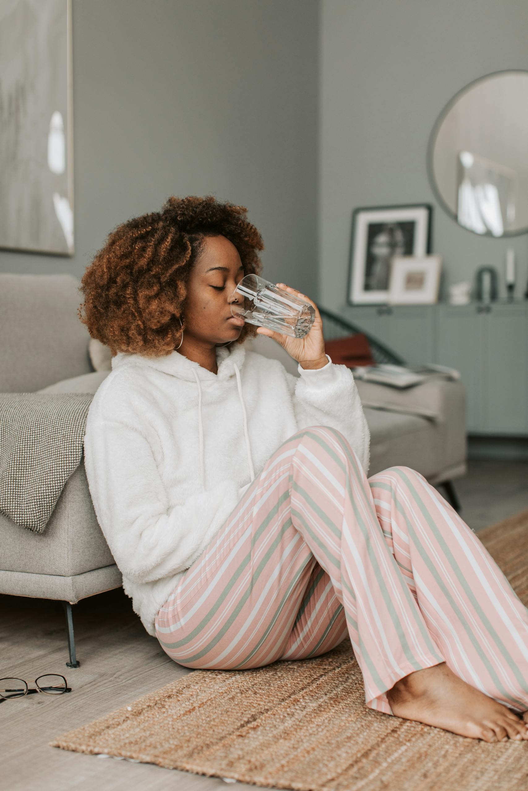 African American woman in hoodie sipping water, sitting on the floor in a cozy home environment.