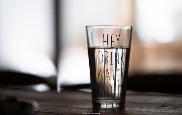 Close-up of a water glass with motivational text 'Hey Drink Water More' in a blurred indoor setting.