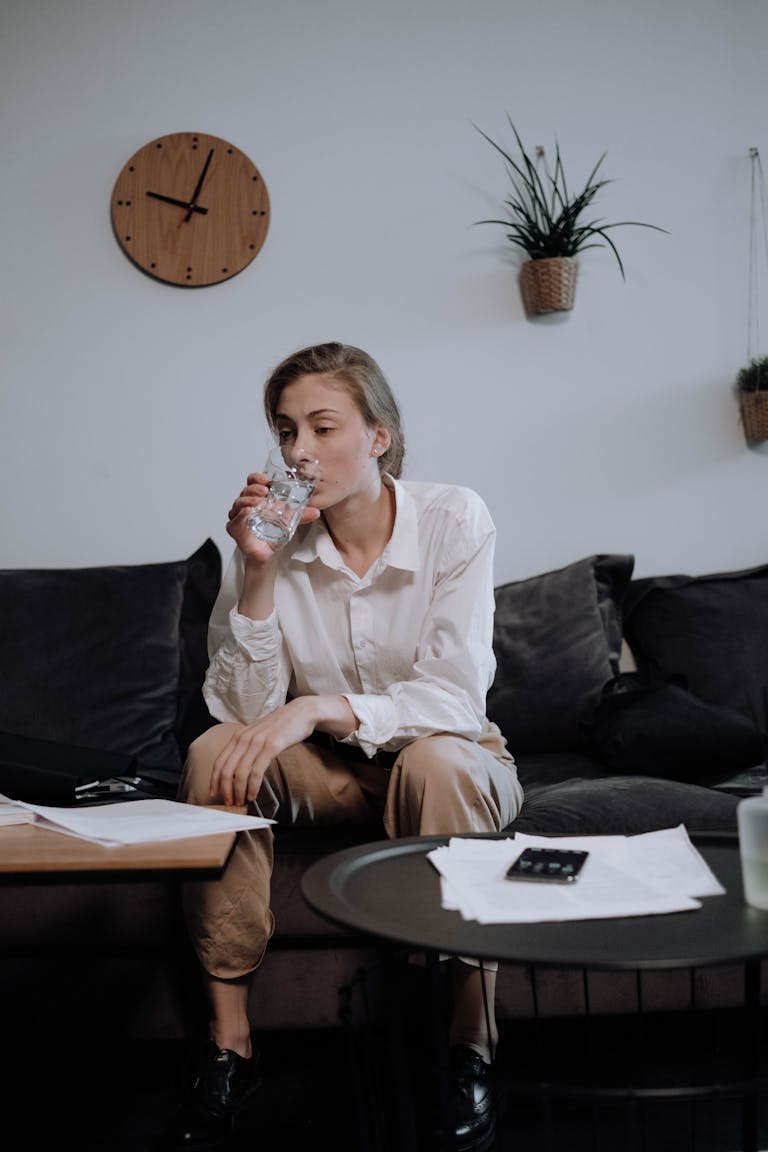Woman in white shirt sitting on a sofa, drinking water and working from home, surrounded by documents.