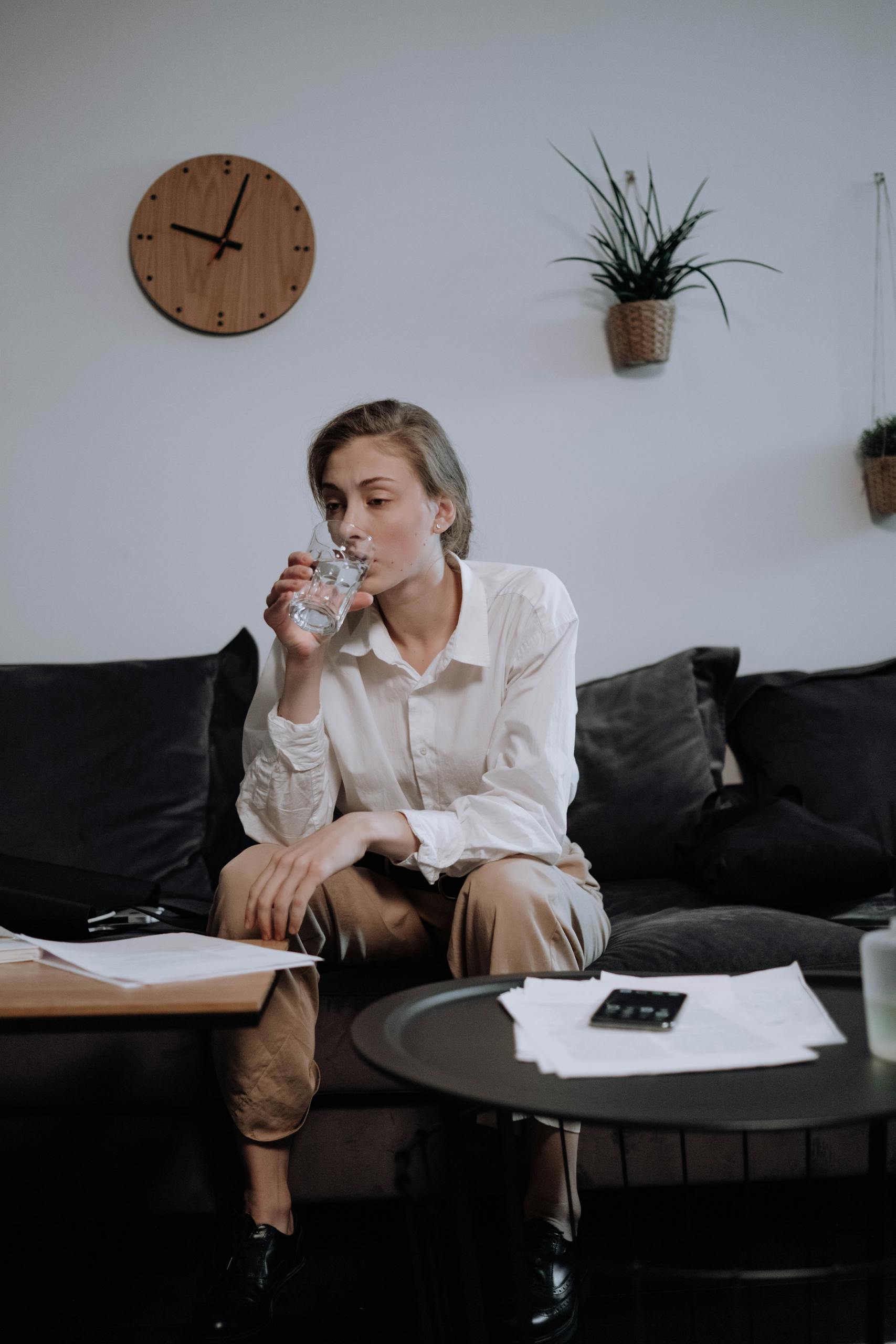 Woman in white shirt sitting on a sofa, drinking water and working from home, surrounded by documents.